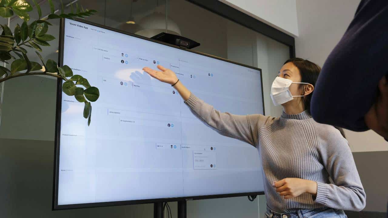 A woman with a face mask presenting a project on a large TV